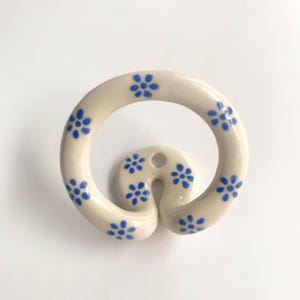 Könnte beinhalten: Ein Wandhaken aus Keramik mit kreisförmigem Schlaufendesign, bestehend aus einer cremefarbenen Basis, die mit kleinen, handgemalten blauen Blumenmustern verziert ist. Der Haken hat ein einzelnes Befestigungsloch oben und eine glatte, glänzende Oberfläche.