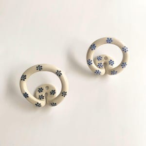 May include: Two circular wall hooks made of off-white ceramic, each featuring a repeating pattern of small blue flowers. The hooks have a rounded, continuous loop design with a central mounting point that includes a small hole for installation.