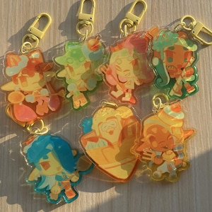 以下が含まれることがあります： 黄色いプラスチック製のロブスタークラスプが付いた、半透明のアクリルキーホルダー7個セット。カラフルな漫画のキャラクターや食べ物をモチーフにしたデザインで、オレンジ、青、緑、黄色などの鮮やかな色合いが特徴です。それぞれのチャームには、キャラクターのイラストが描かれています。
