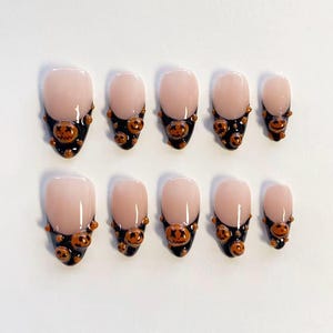 May include: A set of ten press-on nails featuring a nude base with black tips. Each nail is decorated with small, three-dimensional orange pumpkin charms with carved faces, arranged along the black tips of the nails in various sizes.