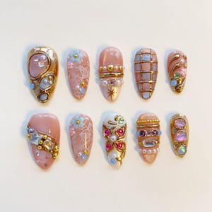 Puede incluir: Un conjunto de diez uñas postizas con diseños variados que incluyen bases rosas, acentos metálicos dorados, perlas incrustadas, cristales transparentes, patrones florales, texturas de encaje y motivos de cuadrícula. Cada uña muestra adornos 3D.