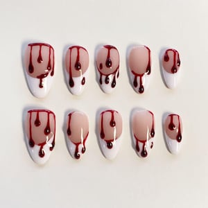 Può includere: Un set di dieci unghie finte a forma di mandorla con base color carne, punte bianche e un design a gocce di sangue rosso lucido. Le unghie sono disposte in due file da cinque, mostrando un motivo coerente di gocce rosso scuro che colano dalle punte bianche verso la base color carne.