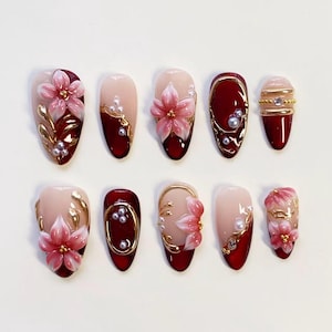 Può includere: Un set di dieci unghie finte caratterizzate da una palette di colori bordeaux intenso e nude. I design includono accenti floreali rosa tridimensionali, dettagli metallici dorati e piccole perle bianche disposte in vari motivi sulle superfici.