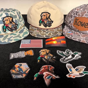 Pode incluir: Três bonés de beisebol com padrões de camuflagem e emblemas de cães com patos, juntamente com uma coleção de emblemas bordados, incluindo uma bandeira americana, uma cena de veado ao pôr do sol e vários desenhos de patos em voo.