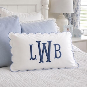 May include: A rectangular white throw pillow featuring a scalloped edge trimmed in light blue piping. The center of the pillow displays a large, embroidered monogram in dark blue thread with the letters L, W, and B in a classic serif font.