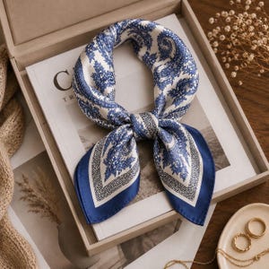 May include: A square silk scarf featuring a blue and white paisley pattern with a dark blue border. The scarf is tied in a knot and displays intricate floral and teardrop motifs throughout the fabric, creating a symmetrical design.