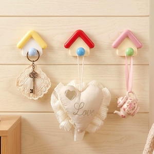 Pode incluir: Três ganchos de parede em forma de casa nas cores amarela, vermelha e rosa. O gancho amarelo segura uma chave num tapete de renda, o vermelho uma almofada em forma de coração com a palavra Love, e o rosa um pequeno ornamento de bule floral.