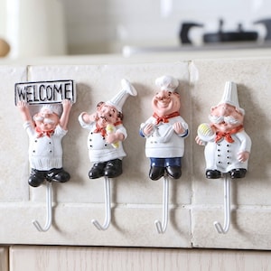 Peut inclure: Quatre crochets muraux représentant des figurines de chefs en uniforme blanc avec des toques et des foulards rouges. Le premier chef tient un panneau avec l'inscription WELCOME. Chaque figurine est fixée à un crochet en métal blanc.