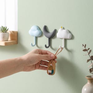 Puede incluir: Tres ganchos de pared con forma de nube azul claro, luna creciente gris oscuro y nube blanca con un pequeño detalle amarillo. Un llavero de madera con un personaje pintado cuelga del gancho de la nube blanca en la pared.