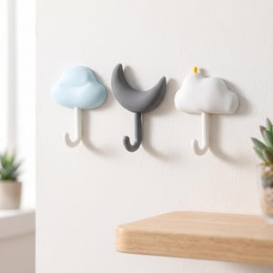 May include: Three wall-mounted hooks featuring a light blue cloud, a dark gray crescent moon, and a white cloud with a small yellow accent. Each hook has a curved, J-shaped hanger extending downward from the central decorative shape.