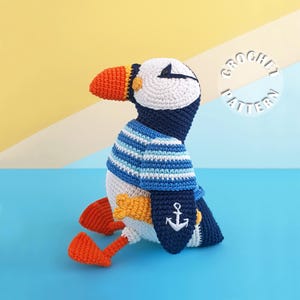 May include: A crocheted puffin toy featuring a white face, a large orange beak, and a dark blue body. The puffin wears a blue and white striped sweater and has a white anchor embroidered on its wing. The text CROCHET PATTERN is visible.