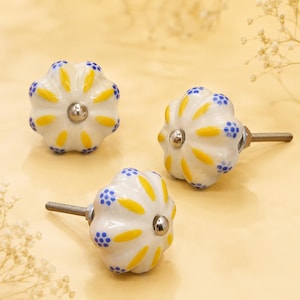 May include: Three white ceramic drawer knobs with a scalloped, pumpkin-like shape. Each knob features a hand-painted pattern of yellow petals radiating from a central silver-toned metal screw, accented by small blue dots along the outer edges.
