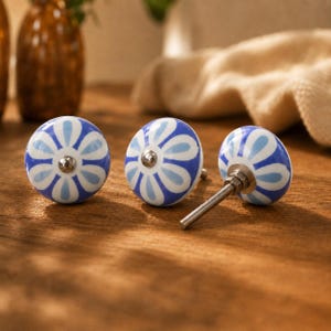May include: Three round ceramic cabinet knobs featuring a hand-painted blue and white floral petal pattern. Each knob has a smooth, glossy finish and a central silver-toned metal screw attachment for installation on furniture or cabinetry doors.