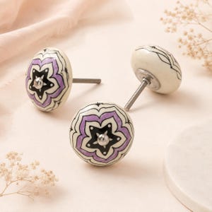 Puede incluir: Dos pomos redondos de cerámica para armarios con base de color crema y un patrón floral pintado a mano en tonos morados y negros. Cada pomo tiene un perno central de metal plateado y un tornillo roscado que sobresale por la parte trasera.