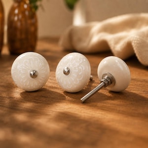 May include: Three round, off-white ceramic cabinet knobs featuring a subtle, embossed floral pattern on the surface. Each knob has a small, centred silver-toned metal accent and a threaded metal bolt extending from the back for installation.
