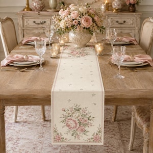 May include: A rectangular table runner featuring a cream-colored fabric with a delicate floral pattern in shades of pink and green. The design includes a large bouquet of roses at the ends and smaller scattered floral motifs along the center.