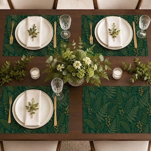 May include: A dining table setting featuring four rectangular placemats with a dark green background and a gold fern leaf pattern. Each place setting includes a white plate, a white cloth napkin topped with a small green sprig, a gold fork, and a gold knife.