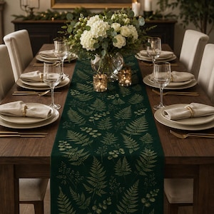 May include: A dark green table runner featuring a repeating pattern of light green fern leaves and eucalyptus sprigs. The runner is centered on a wooden dining table set with white plates, beige napkins with gold rings, and clear glassware.