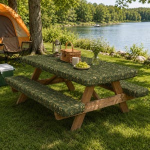 May include: A wooden picnic table set featuring a dark green fabric cover with a small yellow floral pattern on the tabletop and two matching bench cushions. The table is set with a wicker basket, a lantern, a white mug, and a bowl of green grapes.
