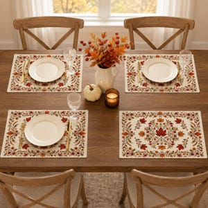 Op de afbeelding: Een set van vier rechthoekige placemats met een crèmekleurige achtergrond en een rand van kleurrijke herfstbladeren, bessen en eikels. Elke placemat is gedekt met een wit keramieken bord, een gouden vork, een gouden mes en een glazen beker.