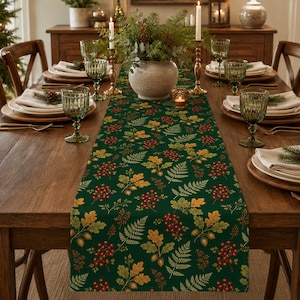 May include: A dark green table runner featuring a repeating pattern of autumn leaves, fern fronds, acorns, and clusters of red berries. The fabric runner is laid flat across a wooden dining table, centered beneath a central floral centerpiece.
