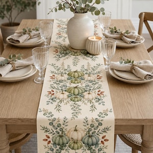 May include: A cream-coloured table runner featuring a repeating pattern of sage green and white pumpkins surrounded by eucalyptus leaves and small berries. The runner is centred on a wooden dining table with place settings and a central vase.