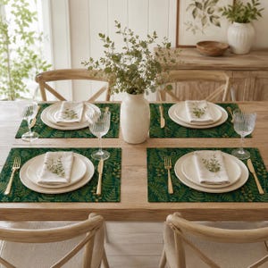 May include: A wooden dining table set for four with dark green fern-patterned placemats, white ceramic plates, folded white napkins, gold-toned flatware, clear ribbed glassware, and a central white vase filled with green leafy branches.