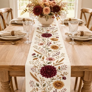 May include: A cream-colored fabric table runner featuring a printed botanical pattern of dark red dahlias, golden yellow chrysanthemums, wheat stalks, and small green leaves, centered on a wooden dining table with place settings and a floral centerpiece.