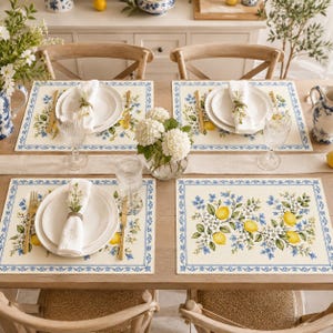 Può includere: Un set di quattro tovagliette rettangolari con sfondo color crema, bordo decorativo blu e una stampa botanica centrale di limoni gialli, fiori bianchi e piccoli fiori blu, disposte su un tavolo da pranzo in legno con piatti bianchi.