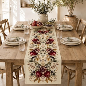 May include: A cream-colored table runner featuring a repeating botanical print of deep red pomegranates, green olive branches, and golden-brown autumn leaves. The runner is centered on a wooden dining table set with white plates and clear glasses.
