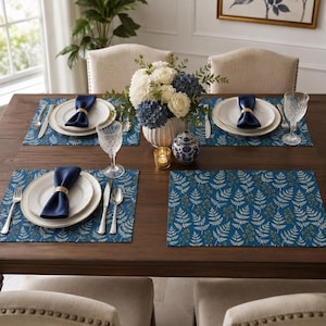 Peut inclure: Une table à manger en bois foncé dressée pour quatre personnes avec des sets de table bleus à motif de fougères blanches. Chaque couvert comprend une assiette blanche, une serviette bleu marine avec un rond de serviette doré, des couverts et un verre.