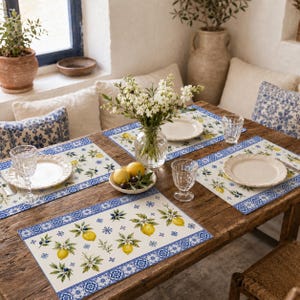 May include: Four rectangular placemats featuring a cream background with a pattern of yellow lemons, green leaves, and white blossoms, bordered by a blue and white geometric tile design. Each placemat is set with a white plate and a clear glass.