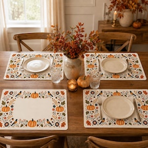 Op de afbeelding: Een set van vier rechthoekige placemats met een crèmekleurig midden en een rand versierd met oranje pompoenen, donkerblauwe vogels, kleurrijke herfstbladeren en eikels. Elke placemat ligt op een houten tafel met een wit bord.