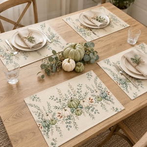 May include: A set of rectangular placemats featuring a printed pattern of sage green pumpkins and eucalyptus leaves on a light beige fabric. Each place setting includes a white ceramic plate, a folded linen napkin, and a clear glass tumbler.