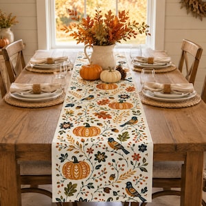 May include: A cream-coloured table runner featuring a repeating pattern of orange pumpkins, blue birds, acorns, and autumn leaves. The runner is centred on a wooden dining table with place settings, a white pitcher, and two pumpkins.