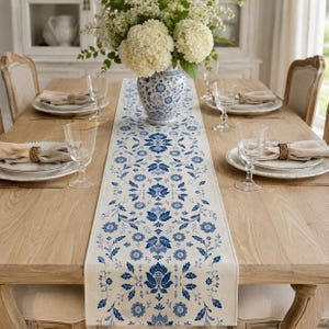 Può includere: Un lungo runner da tavola rettangolare con un motivo floreale blu e bianco al centro di un tavolo da pranzo in legno. Il runner è decorato con intricati motivi botanici ed è accompagnato da piatti bianchi, bicchieri e tovaglioli.