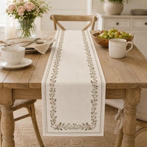 May include: A rectangular table runner made of off-white fabric featuring a delicate botanical border pattern of small pink flowers and green leaves. The runner is centered on a wooden table, framed by a thin green line along its edges.