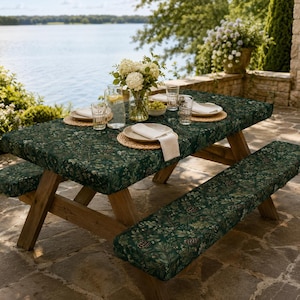 May include: A wooden picnic table and bench set covered in dark green fabric featuring a botanical print of pinecones and foliage. The table is set with white plates, napkins, and clear glasses, with a glass pitcher and a vase of white flowers.
