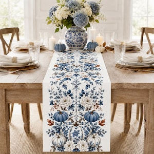 Könnte beinhalten: Ein rechteckiger Tischläufer mit einem sich wiederholenden Muster aus blauen und weißen Kürbissen, Blumenarrangements und Herbstblättern. Das Design ist auf einen weißen Stoff gedruckt und zeigt ein symmetrisches, botanisches Layout.