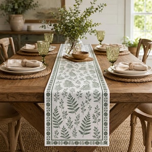 May include: A rectangular table runner featuring a white fabric base with a repeating dark green fern leaf pattern and a floral border. The runner is centered on a wooden dining table set with woven placemats, white plates, and green glassware.