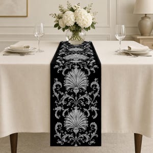 May include: A black table runner featuring a repeating white damask pattern with intricate floral and fan-shaped motifs. The runner is centred on a beige tablecloth, flanked by two clear wine glasses and two white plates with folded napkins.