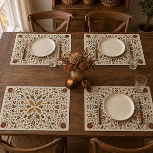 May include: A set of four rectangular placemats featuring a detailed botanical print with autumn-colored leaves, acorns, and flowers in shades of orange, brown, and green. Each placemat is arranged on a wooden table with a ceramic plate, a fork, a knife, and a glass.