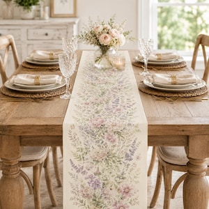 May include: A long, cream-colored table runner featuring a delicate, repeating botanical print of soft pink, lavender, and green wildflowers. The runner is centered on a rustic wooden dining table set with white plates, napkins, and glassware.