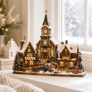 May include: A miniature Christmas village display featuring a central stone church with a clock tower, flanked by a toy shop and a festive building. The scene includes snow-dusted roofs, illuminated windows, a small bridge, a sleigh, and a red mailbox.