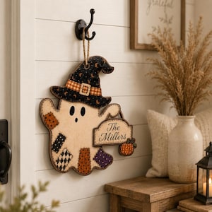 May include: A hanging wooden ghost decoration featuring a black witch hat with orange moon and star patterns, a plaid band, and patchwork-style fabric accents. The ghost holds a sign that reads The Millers and a small orange pumpkin.