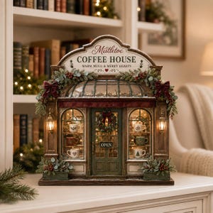 May include: A decorative miniature coffee house building with a sign reading Mistletoe COFFEE HOUSE WARM MUGS & MERRY HEARTS. The structure features arched windows displaying small mugs, a central door with an OPEN sign, and festive greenery.