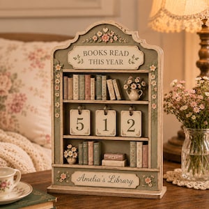 May include: A sage green wooden bookshelf display with the text BOOKS READ THIS YEAR and Amelia's Library. It features miniature book spines, floral accents, and three hanging metal rings displaying the number 512 on white rectangular cards.