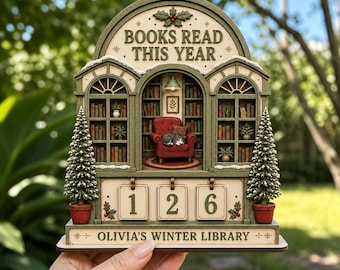 May include: A decorative wooden display titled BOOKS READ THIS YEAR and OLIVIA'S WINTER LIBRARY. It features a miniature library scene with bookshelves, a red armchair, a sleeping cat, and two potted pine trees. Three flip cards show the number 126.