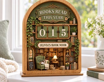 May include: An arched wooden book nook display featuring a flip-style counter showing 015, labeled BOOKS READ THIS YEAR and OLIVIA'S BOOK NOOK. The miniature scene includes a green armchair, a cat, a lamp, and shelves filled with books.