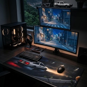 Könnte beinhalten: Ein Computer-Gaming-Setup mit einem schwarzen Tower-Gehäuse mit oranger Innenbeleuchtung, einer mechanischen Tastatur, einer schwarzen Maus und einer großen Schreibtischunterlage mit einem weißen Auto. Zwei übereinanderliegende Monitore.